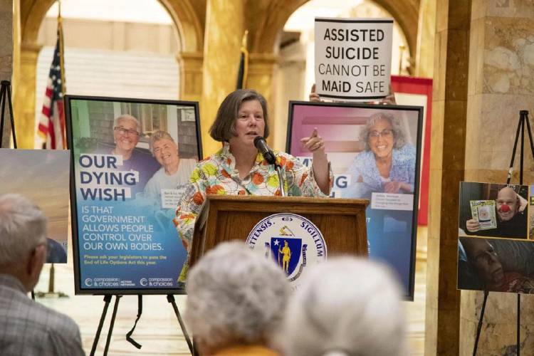 State Sen. Jo Comerford speaks at an event in support of medical aid in dying legislation, while an opponent of the controversial policy holds up a sign behind the podium on Tuesday. 
