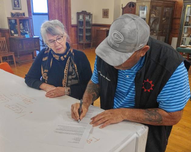 Barre Museum Association board member Elizabeth Martin looks on as Justin Pourier, Fifth Member and Tribal Historic Preservation Officer of the Oglala Sioux Tribe, signs the agreement between the library and the tribe regarding the return of items to the Pine Ridge Reservation. The agreement also carries the signature of board President Ann Meilus. 
